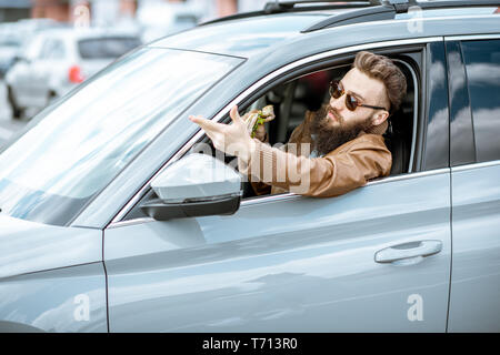 Stilvolle bärtiger Mann einen Snack mit leckeren Sandwich beim Autofahren in der Stadt Stockfoto
