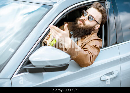 Stilvolle bärtiger Mann einen Snack mit leckeren Sandwich beim Autofahren in der Stadt Stockfoto