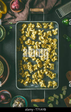 Hausgemachte raw Vegetarische tortellini Pasta in Metall Fach auf dunklen rustikalen Küche Tisch Hintergrund mit Zutaten und Utensilien, Ansicht von oben. Kochen prepar Stockfoto