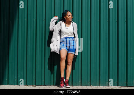 Afrikanische Frau mit dreads Haar, in Jeans Shorts gegen grüne Stahl Wand gestellt. Stockfoto