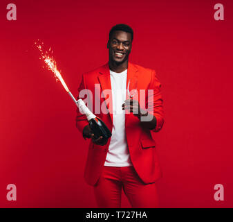 Hübscher junger afrikanischer Mann mit Champagner Flasche, Glas und Feuerwerk in der Hand. Stilvolle Kerl im roten Anzug feiern Silvester vor rotem Hintergrund Stockfoto
