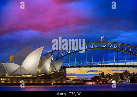 Welt berühmten Sydney Opera House und die Harbour Bridge bei Sonnenuntergang. Unscharfe Wolken und Lichter von Wahrzeichen spiegeln sich in fliessend Wasser des Hafens. Sydney, New Stockfoto