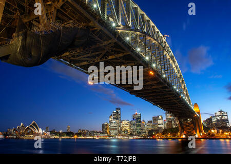 Seitenansicht von der Sydney Harbour Bridge, das Opernhaus und die Innenstadt bei Sonnenuntergang. Beleuchtete Bogen der Brücke in verschwommen Wasser Sydney, New South W widerspiegelt Stockfoto