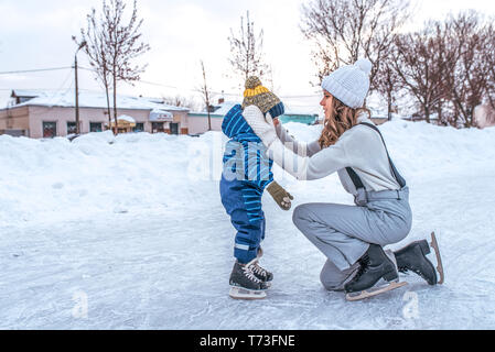 Eine junge Mutter richtet die Junge ihres Sohnes. Im Winter in der Stadt auf der Eisbahn. Im Winter Kleidung Eislauf, auf einem Hintergrund von Schnee. Freier Speicherplatz für Stockfoto