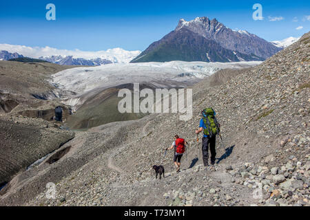 Das Paar und ihr Hund sind auf der Gletschermorraine unterwegs, auf dem Weg zum Root Glacier, mit dem Donoho Peak und den Wrangell Mountains im Hintergrund... Stockfoto