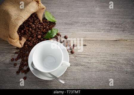 Leere weiße Tasse und Untertasse mit frisch gerösteten Kaffee Bohnen verschütten aus einem kleinen hessischen Tasche gesehen von oben nach unten auf rustikalen verwittertes Holz mit Kopie Raum Stockfoto