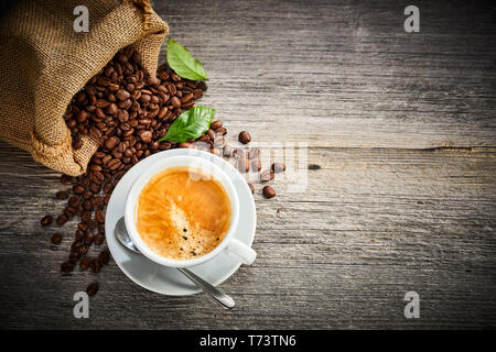 Tasse frisch gebrühten heißen schaumigen Espresso Kaffee mit gerösteten Bohnen Verschütten von einem hessischen Tasche auf rustikalem Holz am besten mit Kopie Raum und v Stockfoto