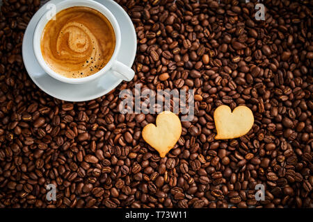 Zwei herzförmige Plätzchen mit Tasse heißen schaumigen Espresso auf einem Hintergrund von gerösteten Kaffeebohnen braun Gesehen von oben nach unten in einem konzeptionellen Bild Stockfoto