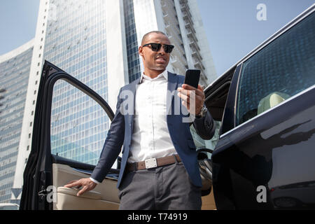 Junge schwarze Unternehmer tragen Sonnenbrillen gehen aus dem Auto halten Sie das Smartphone auf Bildschirm schaut besorgt verpasster Anruf Stockfoto