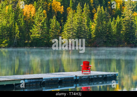 Muskoka Chair auf Dock mit Herbst bunte Laub in der ruhigen See Wasser von Clear Lake, Riding Mountain National Park; Manitoba, Kanada wider Stockfoto