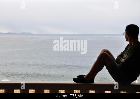 Ein Mann sitzt auf dem Geländer und Blick auf das Meer, Silhouette. Stockfoto