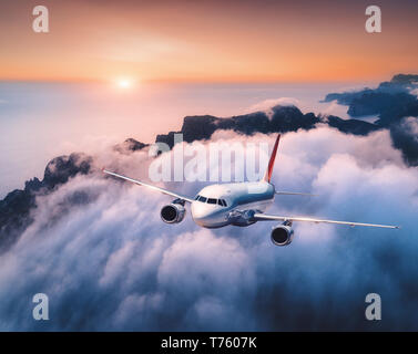 Pkw Flugzeug fliegen über Wolken bei Sonnenuntergang. Landschaft mit weißen Flugzeug, niedrige Wolken, Meer, orange sky in der Abenddämmerung. Flugzeug ist. Bu Stockfoto