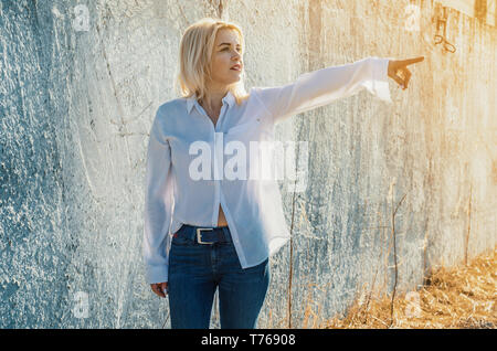 Attraktive Frau im weißen Hemd steht auf dem Hintergrund einer konkreten Zaun, zeigt mit dem Finger auf der rechten Seite. Stockfoto