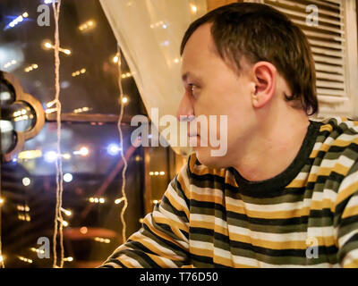 Ein einsamer Mann im gestreiften Pullover sitzt abends in einem Cafe, schaut aus dem Fenster Stockfoto