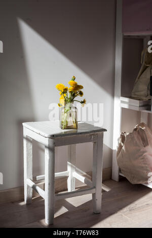 Ein Strauß gelber Löwenzahn in einem Glas transparent Glas auf ein weißes Holz Hocker vor dem Hintergrund der Wand Stockfoto