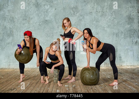 Eine Gruppe von schönen Fitness Mädchen in ein Fitnesscenter in der Nähe einer grauen Wand mit Requisiten für Training, emotional, am Telefon zu sprechen. Kopieren Sie Raum, grau Stockfoto