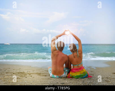 Junges Paar in Liebe am Strand zu sitzen und faltete seine Hände in die Form eines Herzens, durch die die Sonne scheint. Das Konzept der Hochzeitsreise, Liebe Stockfoto