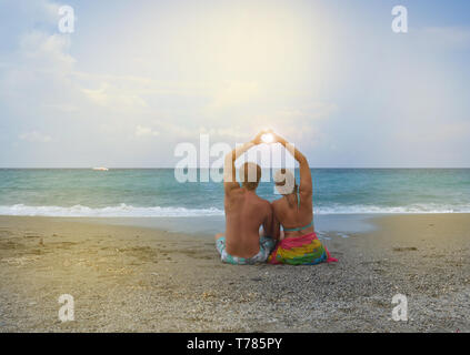 Junges Paar in Liebe am Strand zu sitzen und faltete seine Hände in die Form eines Herzens, durch die die Sonne scheint. Das Konzept der Hochzeitsreise, Liebe Stockfoto