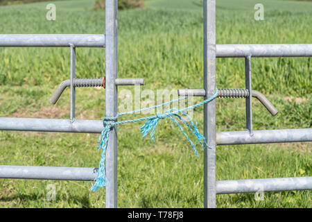 Verzinktes Stahlfarmtor über dem Eingang zum Feld, aber geschlossen mit blauem Polyesterseilknoten. Für den Zugang verweigert, Barriere, halten Sie aus, Gras grüner Idiom Stockfoto