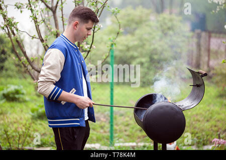 Junger Mann, der in der Nähe von Grill. Das Anzünden von Feuer im Freien. Stattliche männliche Athleten im Hof in der Nähe von Ihrem Haus bereitet Grill outdoo Stockfoto