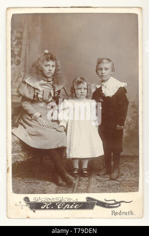 Originale viktorianische Kabinettkarte von 3 gut gekleideten, eleganten kornischen Kindern, Bruder mit Schwestern in hübschen Kleidern, aus den Studios von H. Opie, Redruth, Cornwall, Großbritannien um 1895,1896 Stockfoto