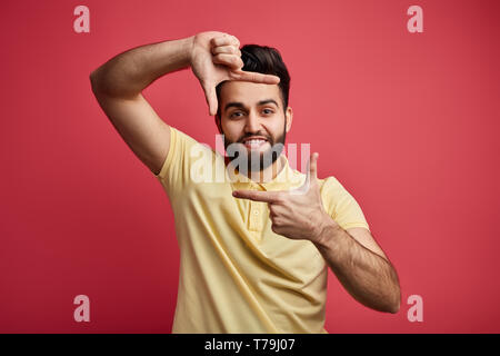Junge positive männliche in gelben T-Shirt, Rahmen mit seinen Händen, in die Kamera schauen und lächeln. close up Portrait. Menschen. Isolierte rosa Hintergrund Stockfoto