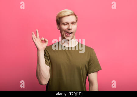 Junge freundliche gerissen charmante Kerl winks und zeigt OK Geste, Stöcke, seine Zunge isoliert auf rosa Hintergrund. bis Foto schliessen. Stockfoto