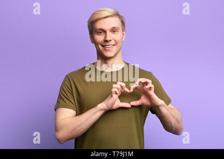 Gerne attraktive Blondine junger Mann, Hände halten im Herzen Geste, seine Emotionen, Liebe, über violett Hintergrund isoliert. Stockfoto