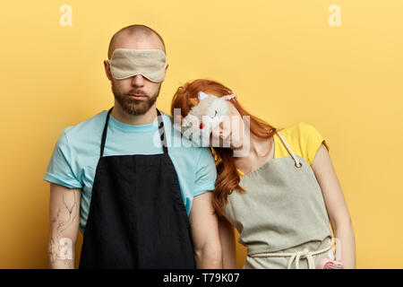 Attraktiver Mann und schöne Ingwer Frau in eine Schürze und Nacht, Schlafmaske mit müde und krank Ausdruck auf dem gelben Hintergrund isoliert. Coup Stockfoto