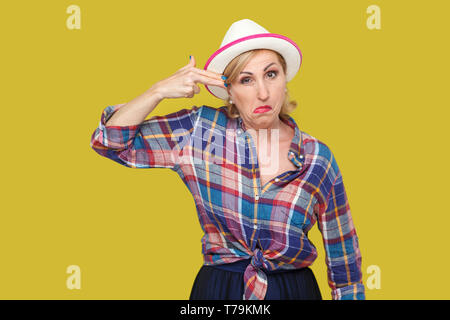 Portrait verrückt der Moderne, elegante, reife Frau im lässigen Stil mit weißen Hut stehen und von der Kamera mit Pistole Pistole Geste. indoor Studio Stockfoto