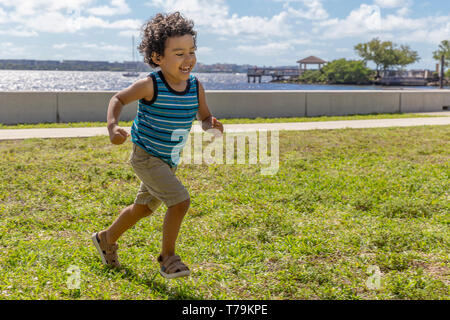 Ein kleiner Junge läuft über das Feld, während laut lachen. Das glückliche Kind, eine wundervolle Zeit im Freien in der Waterfront Park. Stockfoto