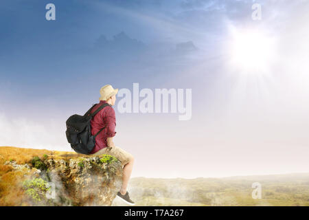 Ansicht der Rückseite des asiatischen reisenden Mann mit Hut und Rucksack sitzt am Rande der Klippe an Landschaften mit Sonnenlicht Hintergrund Stockfoto