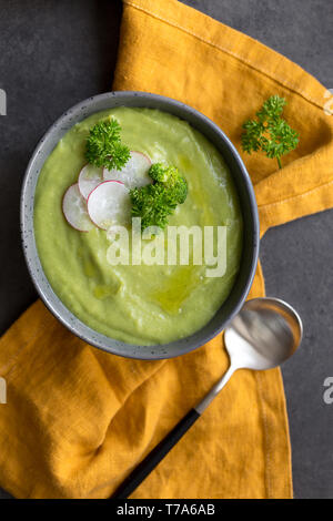 Broccoli creme Suppe mit Radieschen und Löffel auf grauen Platten, mit orange Serviette auf grauen Hintergrund. Frische rohe Brokkoli in der Nähe. Konzept der gesunden Ernährung. Fla Stockfoto