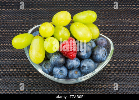 Traube Traube, Blaubeeren, Himbeeren in Glasschale. Süsse saftige Beeren im Detail. Farbige Früchte in ovalen Teller auf schwarz gestreifte Tischset. Gesunden Snack. Stockfoto