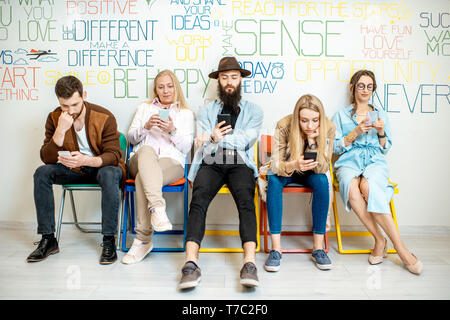 Gruppe von verschiedenen Leuten aus Smartphones beim zusammen in einer Reihe sitzen auf der Wand Hintergrund im Innenbereich Stockfoto