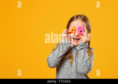 Portrait von ein fröhliches kleines Mädchen Kind auf einem gelben Hintergrund. Schülerin hält die Buchstaben A und B der Begriff der Bildung und Schule. Stockfoto