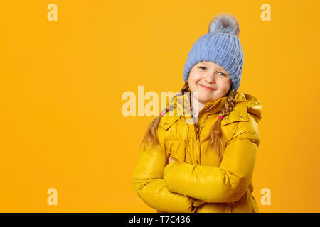 Closeup Portrait von ein fröhliches kleines Mädchen in Jacke und Strickmütze auf gelben Hintergrund. Das Kind faltete ihre Arme und sieht in die Kamera. Stockfoto