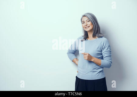 Asiatische Middle-Aged Frau mit grauen Haaren steht auf einem weißen Hintergrund im Studio lächelnd. Geneigter Kopf, eine Hand, eine Hand auf die Taille. Sie trägt Blau Stockfoto