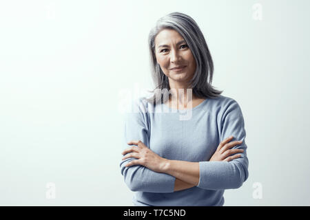 Schöne asiatische Frau mit grauen Haare und dunkle Haut steht auf einem weißen Hintergrund im Studio Bitte Lächeln. Geneigter Kopf, Hände gekreuzt Stockfoto