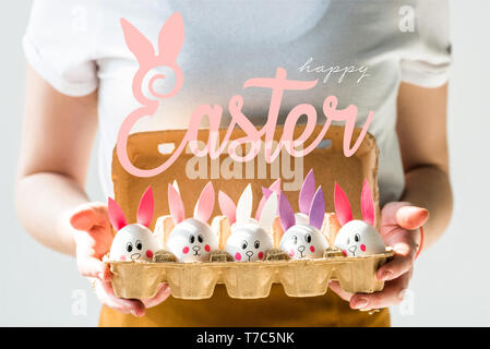 7/8-Ansicht von Frau mit ei Fach mit Huhn Eier mit gezeichnet Kaninchen Gesichter und Papier Ohren und frohe Ostern Schrift Stockfoto