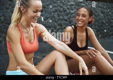 Zwei passen junge Frauen in Sportkleidung in einem Parkplatz außerhalb sprechen und gemeinsam Lachen nach dem Training sitzen Stockfoto