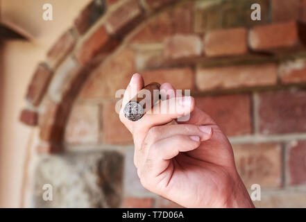 Ein Mann hält eine brennende kubanischen Zigarre in der Hand und Rauchen. Raucher vor Defokussierten Mauer. Stockfoto