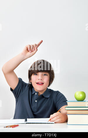 Aufgeregt und Glücklich lächelnde kaukasischen Junge am Schreibtisch anheben Hand mit Zeigefinger sitzen die Frage im Unterricht in der Schule zu beantworten Stockfoto