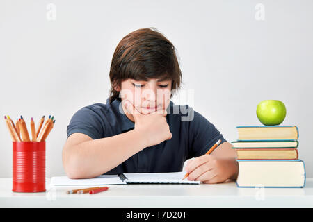Kaukasische Schule Junge sitzt am Tisch und denken mit einem seriösen und konzentrierten Ausdruck, während seine Hausaufgaben machen. Stockfoto