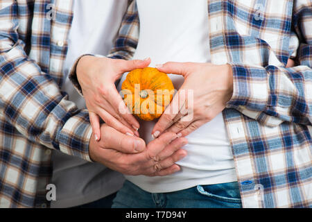 Mann und Frau in Jeans und ein kariertes Hemd hält wenig Kürbis in ihren Händen. Paar ein Herz mit den Fingern auf dem Bauch der werdenden Mutter Stockfoto