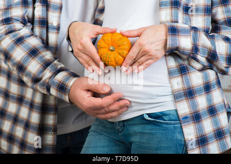 Mann und Frau in Jeans und ein kariertes Hemd hält wenig Kürbis in ihren Händen. Paar ein Herz mit den Fingern auf dem Bauch der werdenden Mutter Stockfoto