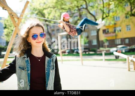 Teen Age Girl mit Sonnenbrille posing und Kind in Rosa cap Schwingen auf Spielplatz im Hintergrund - unbeschwerte Kindheit Stockfoto