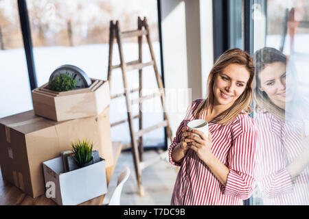 Eine glückliche junge Frau im neuen Haus bewegen, stützte sich auf ein Fenster und einen Kaffee zu trinken. Stockfoto