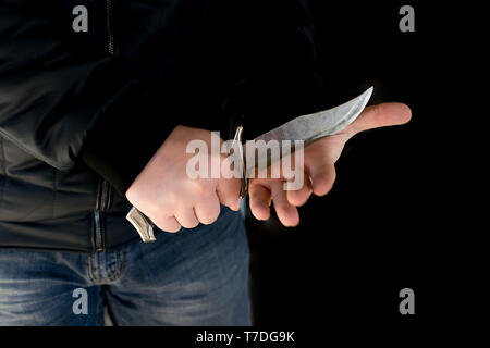 Kriminalität, ein Mann in der Lederjacke und Jeans in einem dunklen Raum trotzig schneidet seine Hand mit einem Messer Stockfoto