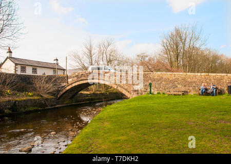 Paar auf einer Bank sitzen durch steinerne Brücke über den Fluss Dunsop auf der Straße in Dunsop Brücke Dorf Trog von Bowland Lancashire England Großbritannien Stockfoto